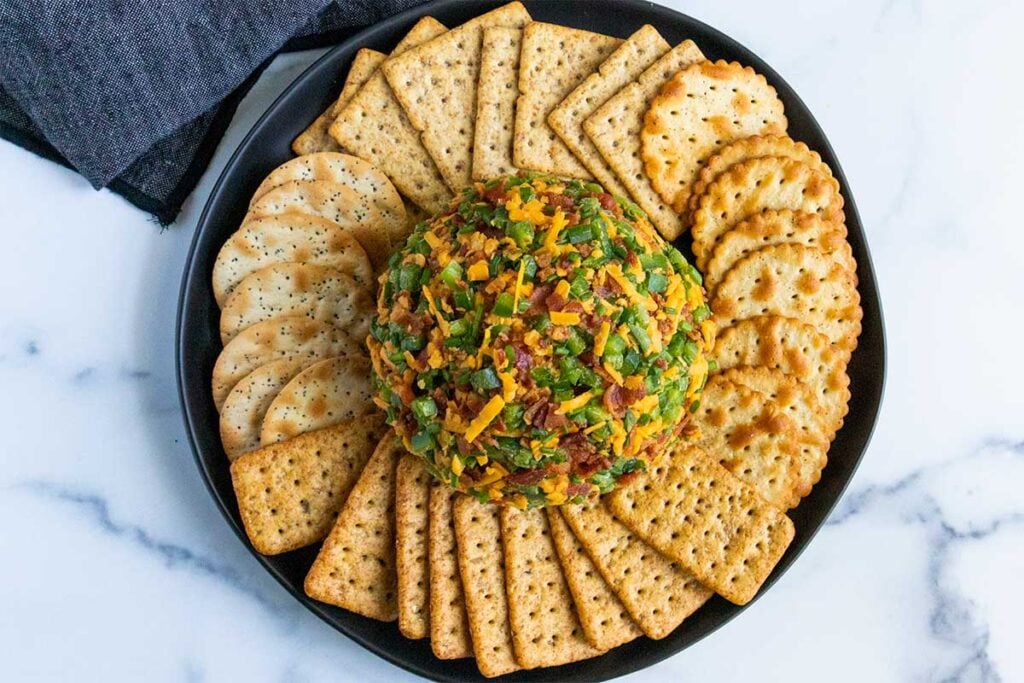 Jalapeno popper cheese ball with bacon and jalapenos rolled onto the surface surrounded by crackers on a dark plate.
