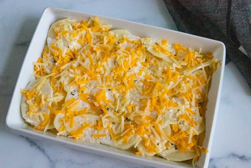 Potatoes au gratin topped with shredded cheddar cheese in white baking dish.
