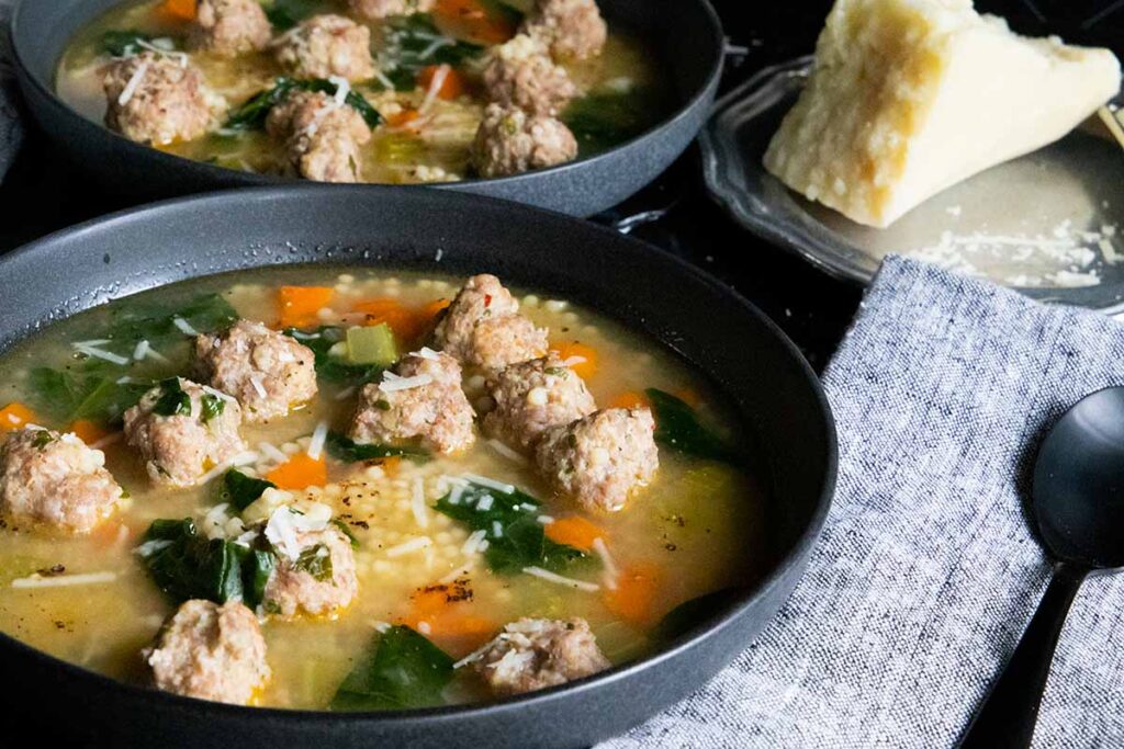 Italian wedding soup in black bowl and a slab of Pecorino Romano on a silver plate.