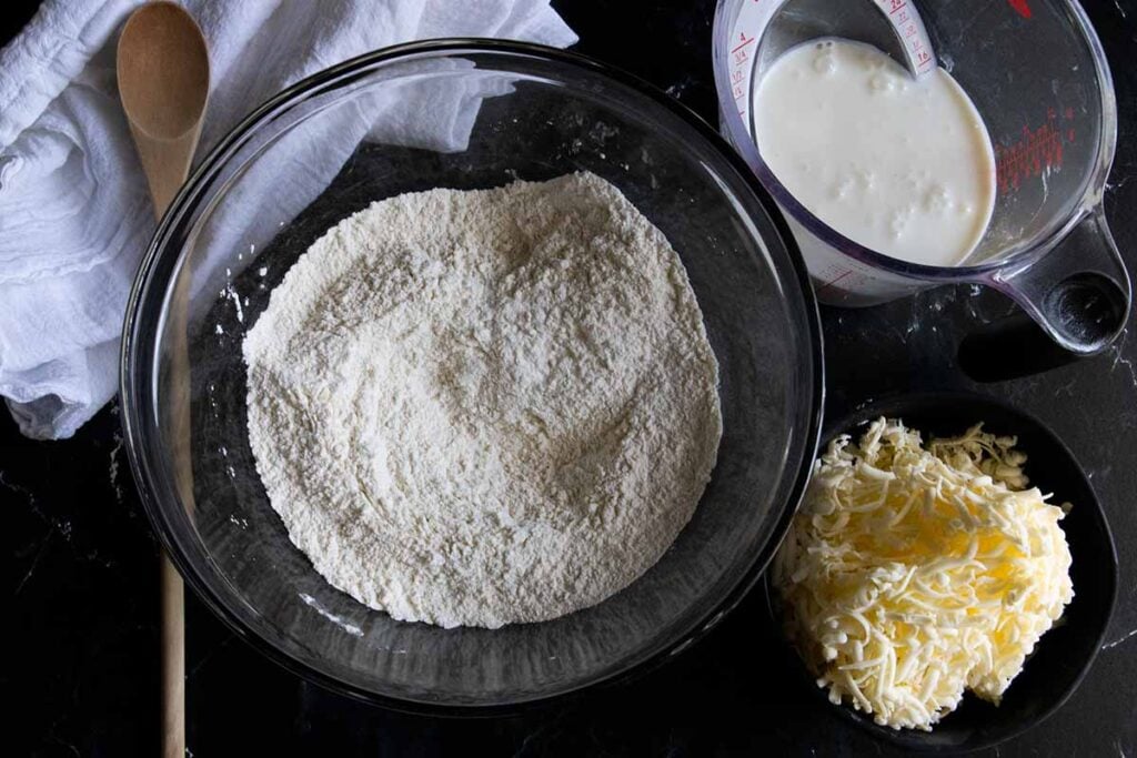 Dry ingredients in a glass mixing bowl, buttermilk in a measuring cup, frozen grated butter in a small black bowl.