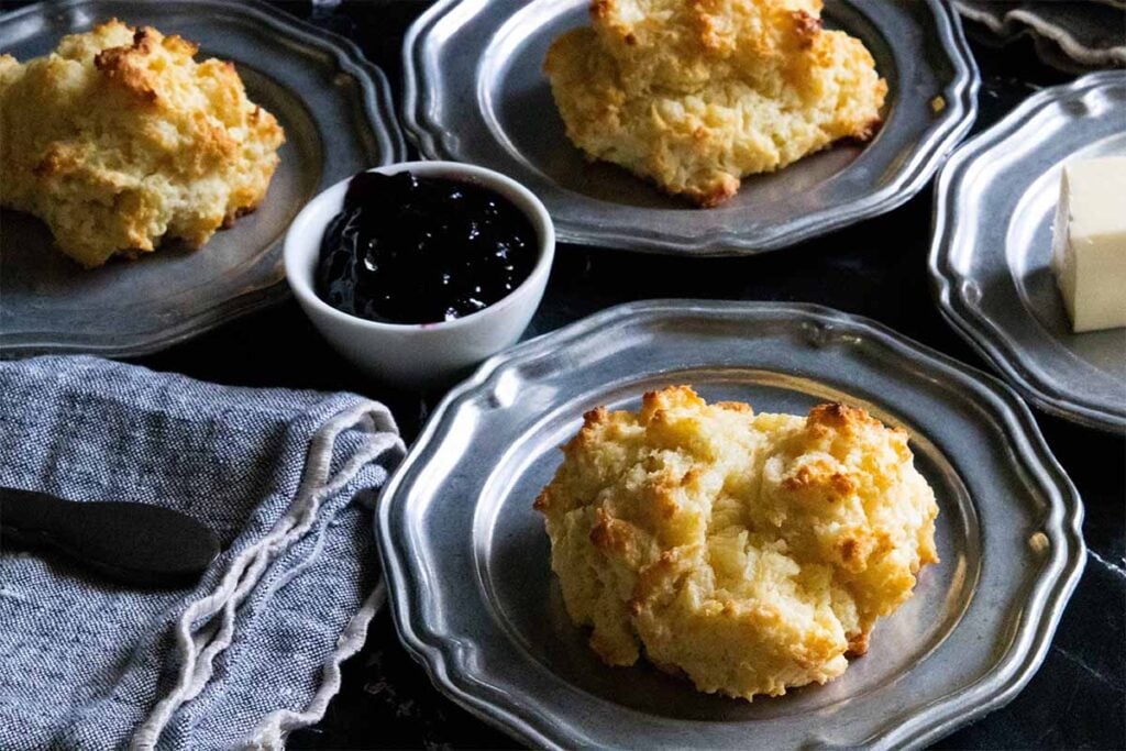 Buttermilk drop biscuits on silver plates with jam in a small bowl, and a butter stick on side plate.
