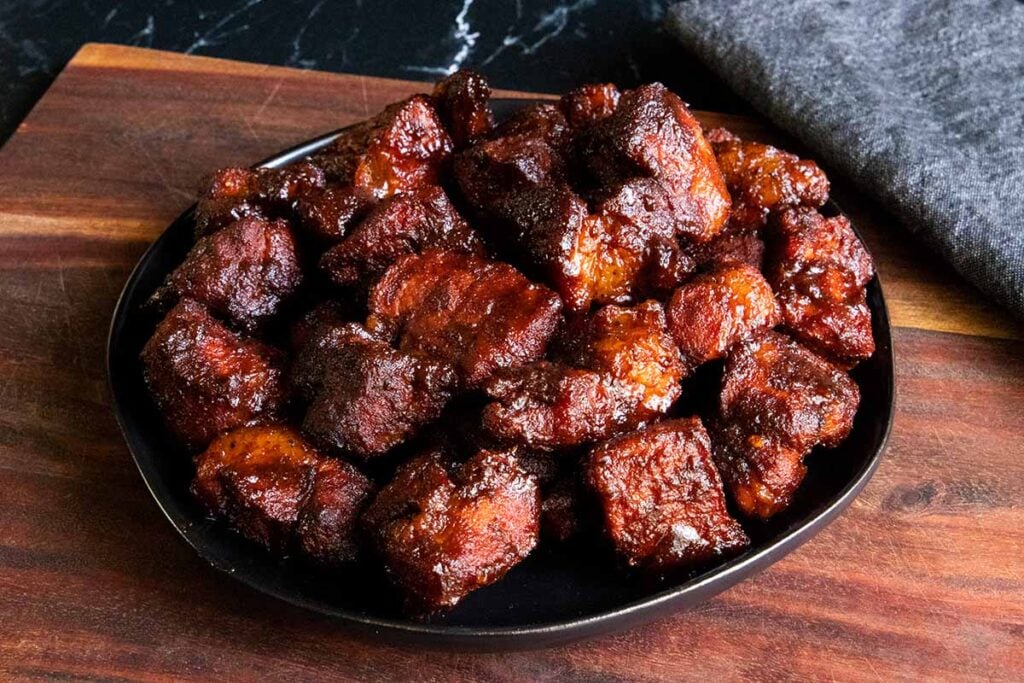 Pork butt burnt ends piled on a black plate on top of wooden board.