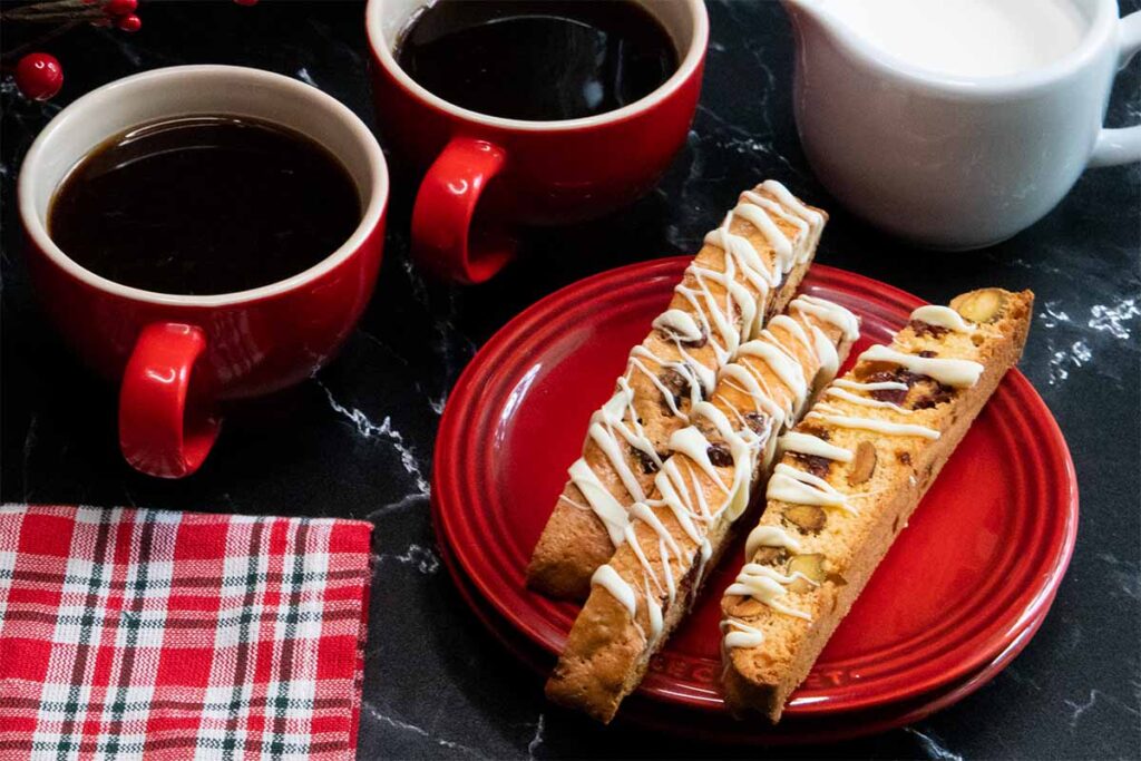 White chocolate cranberry pistachio biscotti on a red plate next to red coffee mugs.