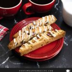 White chocolate cranberry pistachio biscotti on a dark plate next to dark coffee mugs.