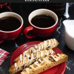 White chocolate cranberry pistachio biscotti on a dark plate next to dark coffee mugs.