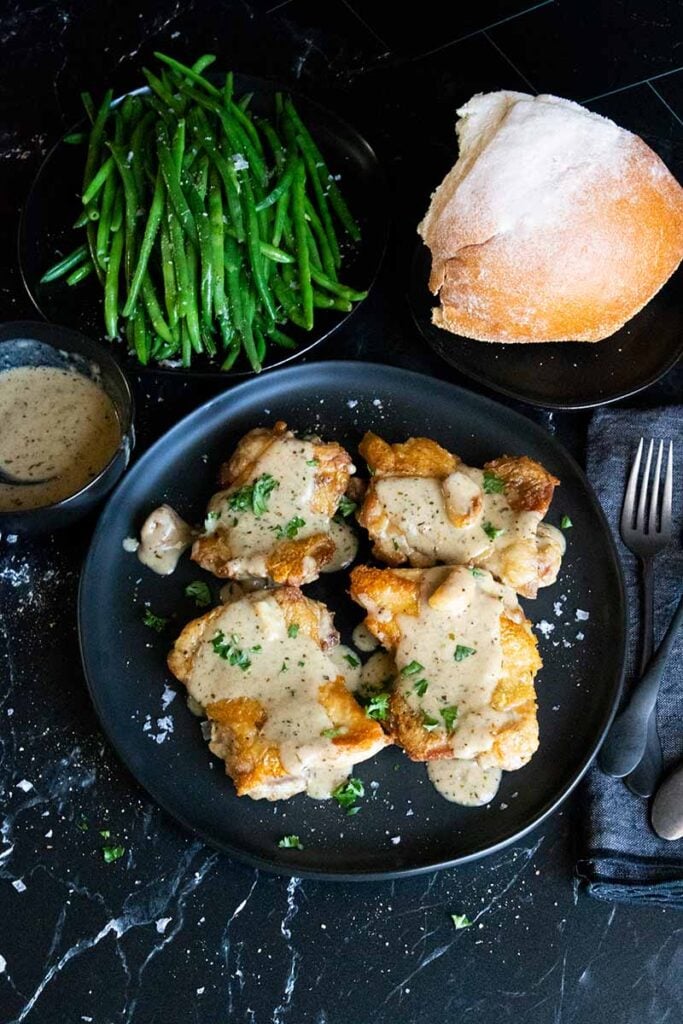 Crispy chicken thighs with a garlic sauce on a black plate. Green beans and bread are in the background.