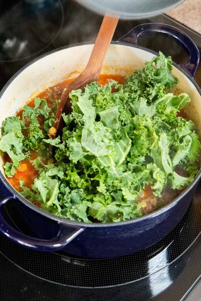 Kale being added to the pot of lentil soup with Italian sausage.