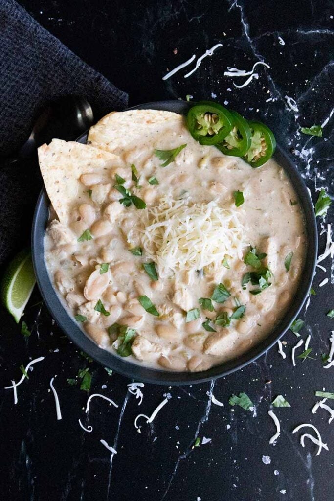 White chicken chili in a black bowl garnished with jalapenos, cilantro, tortilla chips and a lime wedge