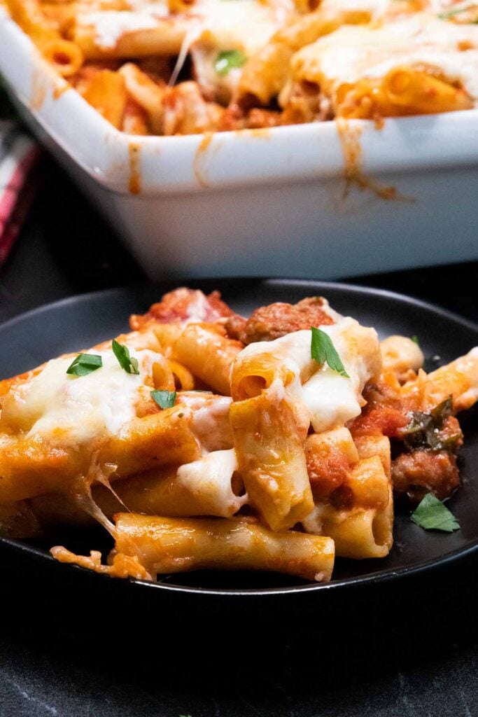A serving of baked ziti on a black plate with a casserole dish in the background.