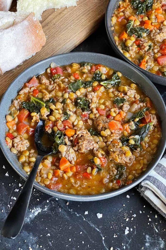 Two bowls of sausage lentil soup and some bread on a cutting board.