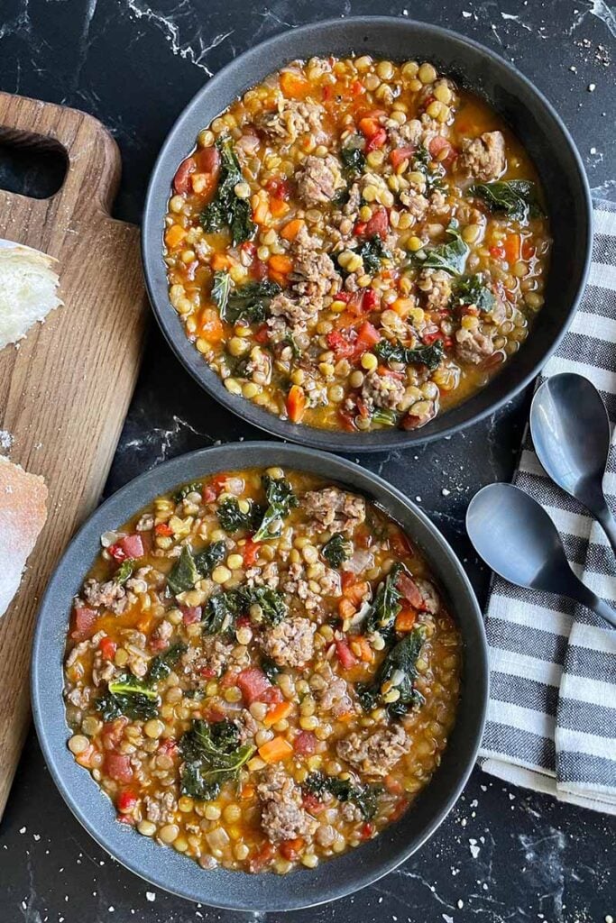 Two bowls of lentil sausage soup in  dark bowls, with bread slices on a wooden board.