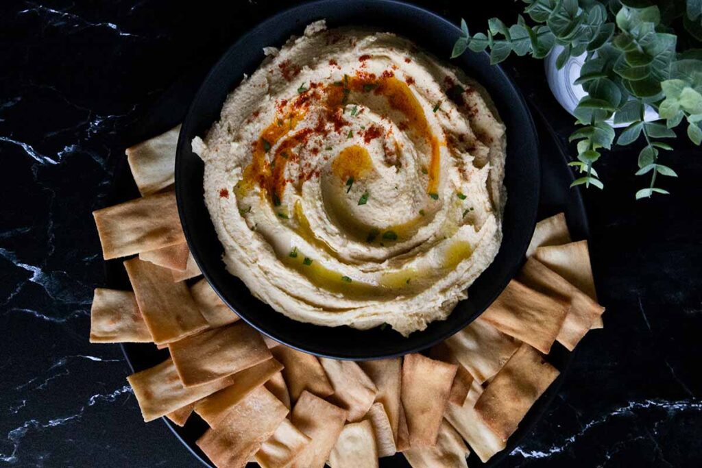 Homemade hummus in a black bowl surrounded by pita chips.