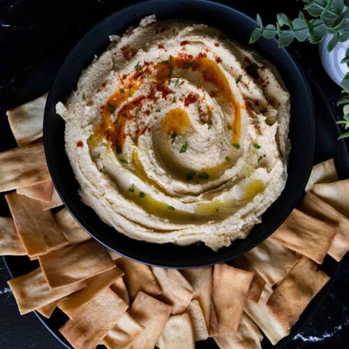 Homemade hummus in a black bowl surrounded by pita chips.