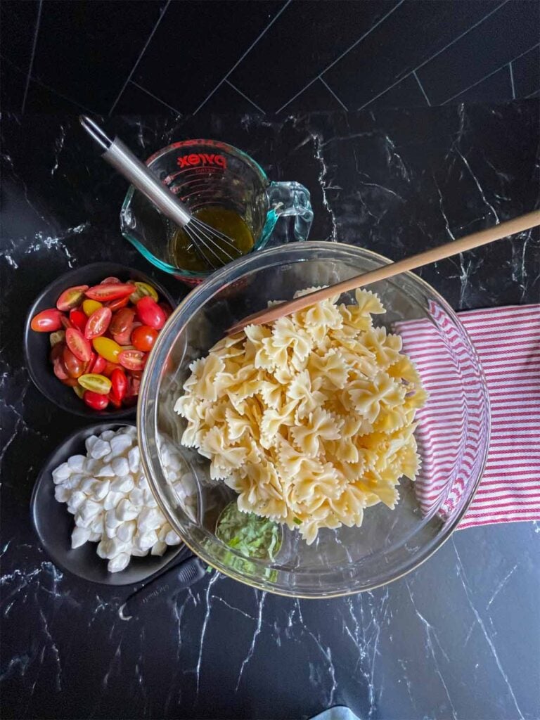 Cooked pasta in a glass mixing bowl, mozzarella in a small black bowl, tomatoes in a small black bowl, and the vinaigrette in a measuring cup.