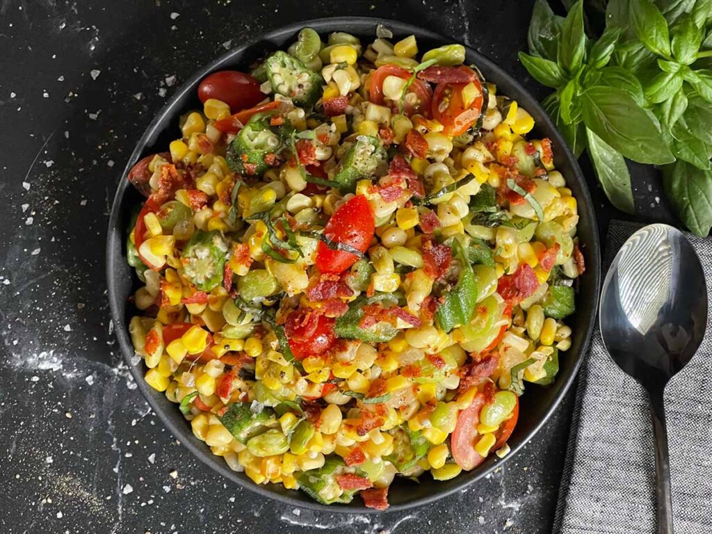 Summer succotash in a black bowl with a bunch of fresh basil in the background.