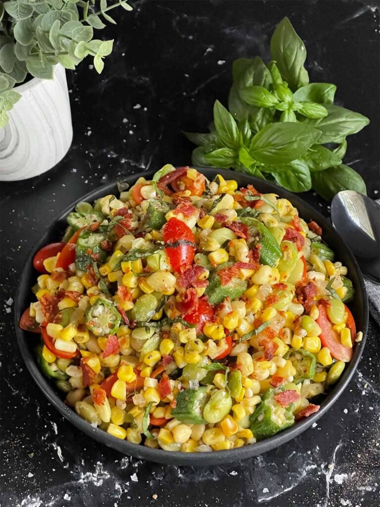 Summer succotash in a black bowl with a bunch of fresh basil behind the bowl.
