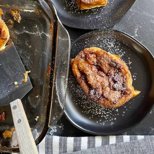 Slices of overnight maple bacon baked french toast on black plates garnished with powdered sugar.