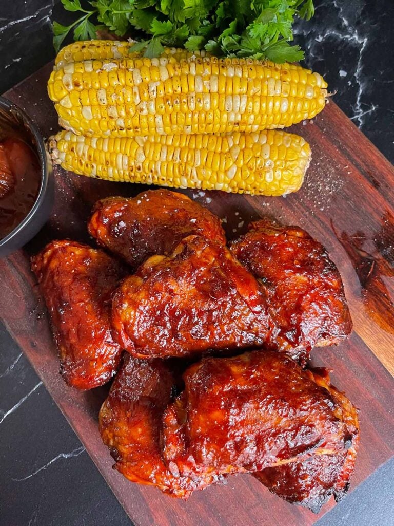 BBQ chicken thighs with crispy skin. Grilled yellow corn as a garnish on a brown cutting board.