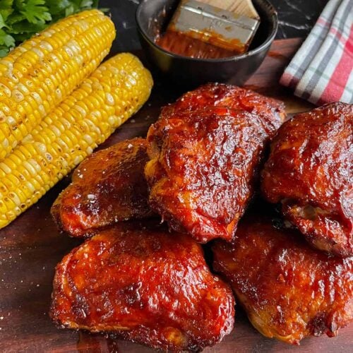BBQ chicken thighs with crispy skin and grilled corn on a cutting board.