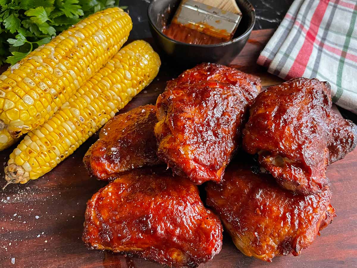 BBQ chicken thighs with crispy skin and grilled corn on a cutting board.