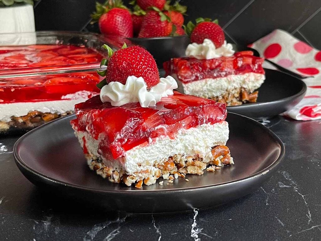 Slice of strawberry pretzel salad on black plates garnished with whipped cream and a fresh strawberry.