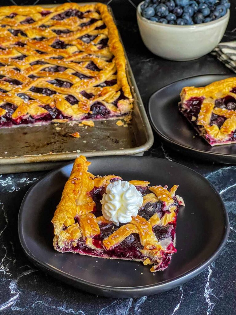 Slice of blueberry slab pie garnished with whipped cream on a black plate.