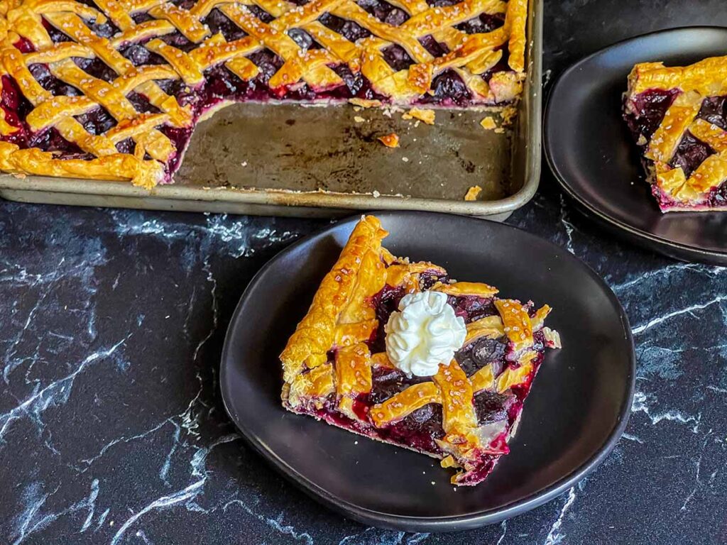 A slice of blueberry slab pie on a black plate with the whole pie in the background.