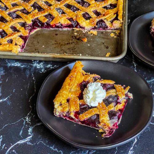 A slice of blueberry slab pie on a black plate with the whole pie in the background.