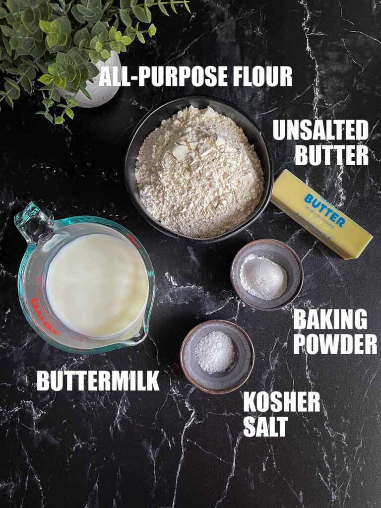 Flour, butter, buttermilk, baking powder, and kosher salt displayed on a black marble counter top.