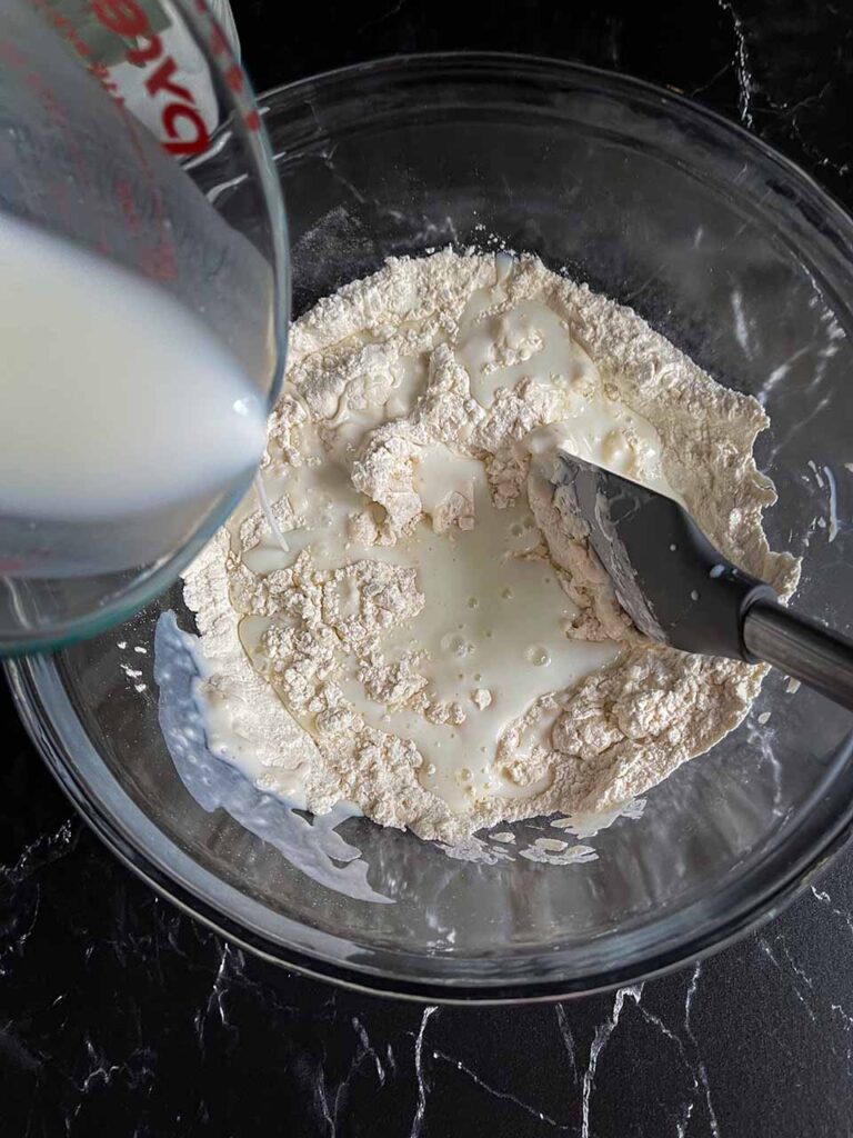 Buttermilk being stirred into the dry ingredients in a glass mixing bowl.