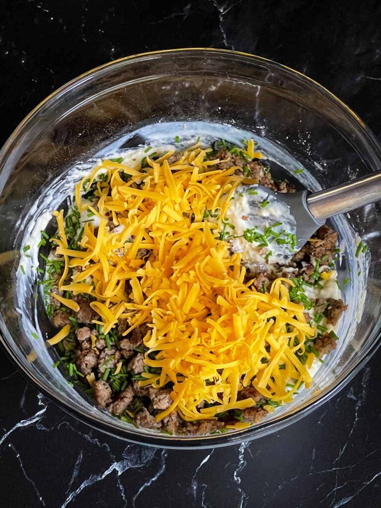 Cooked sausage, shredded cheese, and chives being stirred into the batter with a silver spatula.