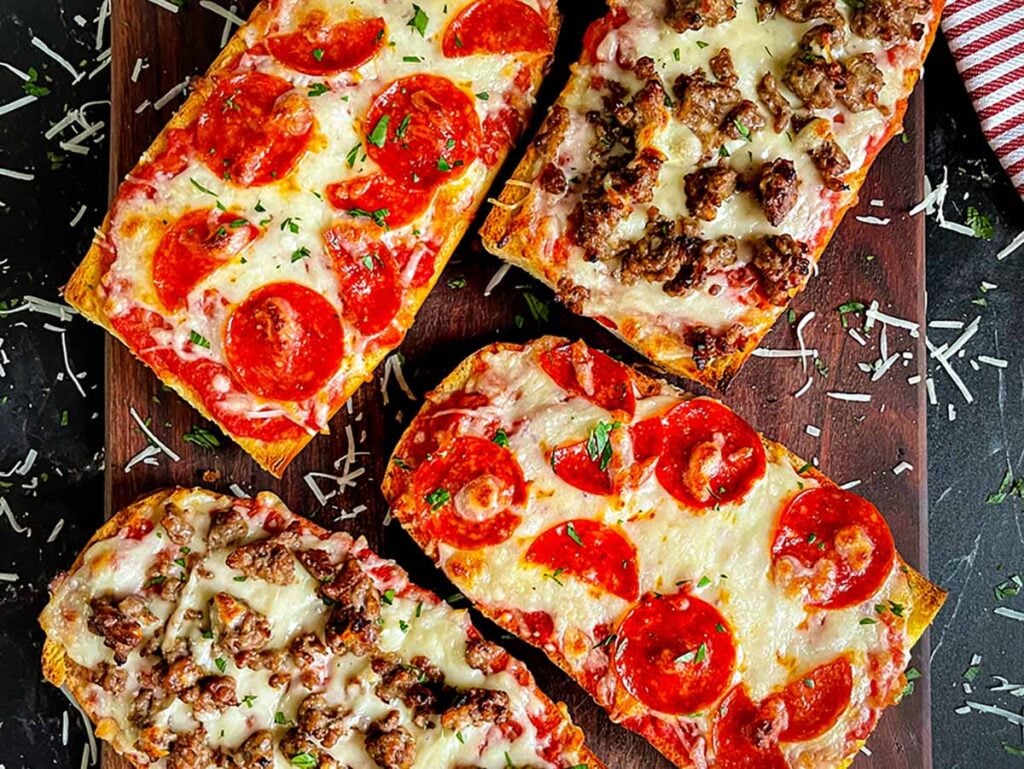 French bread pizza with pepperoni and italian sausage on a dark wooden cutting board.