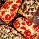 French bread pizza with pepperoni and italian sausage on a dark wooden cutting board.