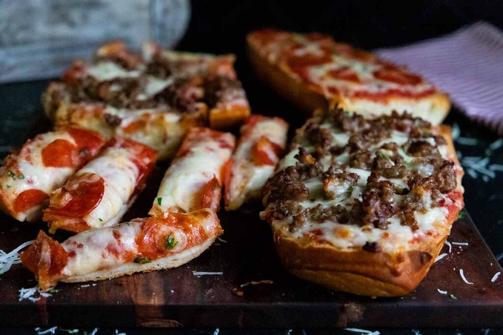 French bread pizzas with pepperoni and italian sausage on a wooden cutting board.