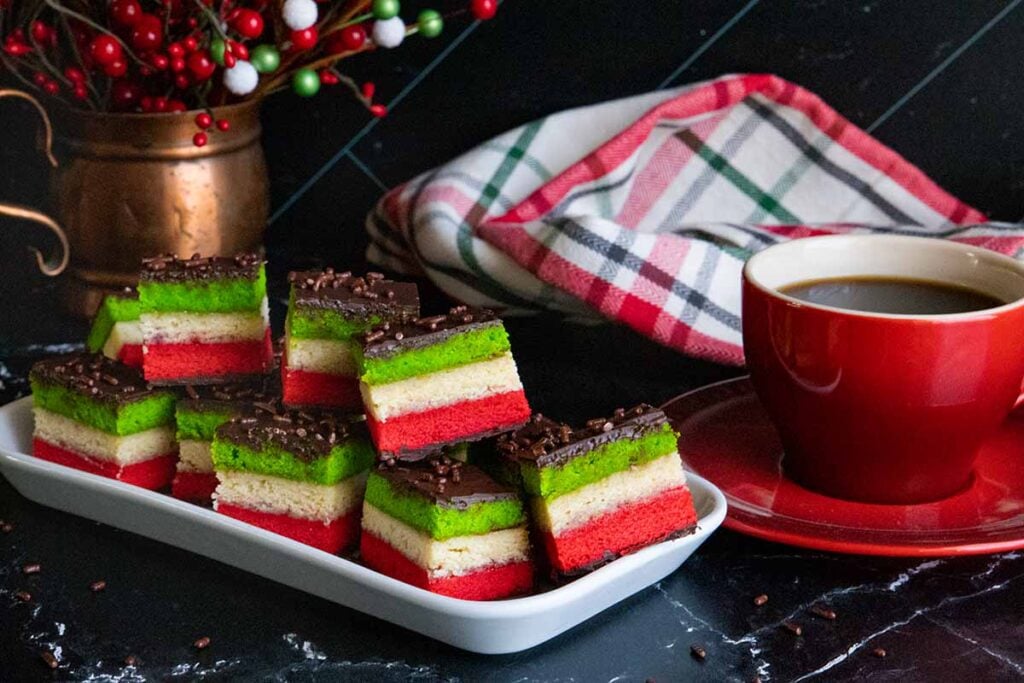 Italian rainbow cookies on a white tray with a red cup of coffee.