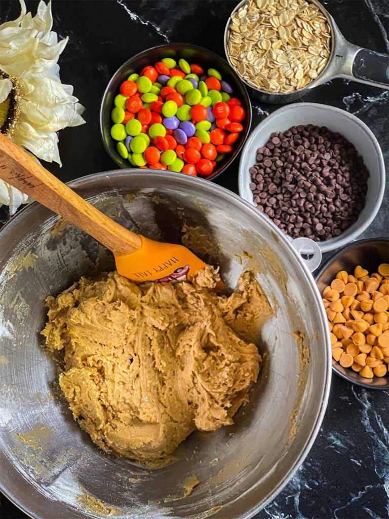 Cookie dough in a metal mixing bowl with the candies and oatmeal on the side.