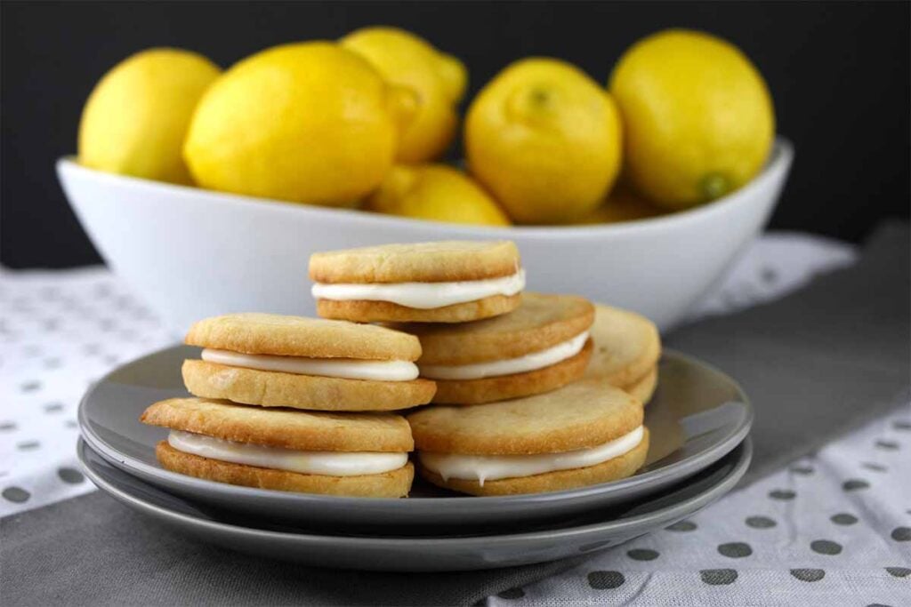 Lemon Sandwich Cookies stacked on gray plate in front of a white bowl of lemons.