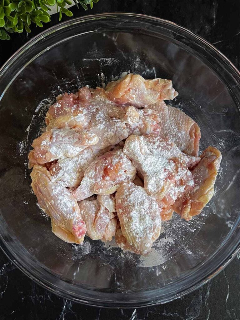 Raw chicken wings in a glass bowl with baking powders sprinkled over them.