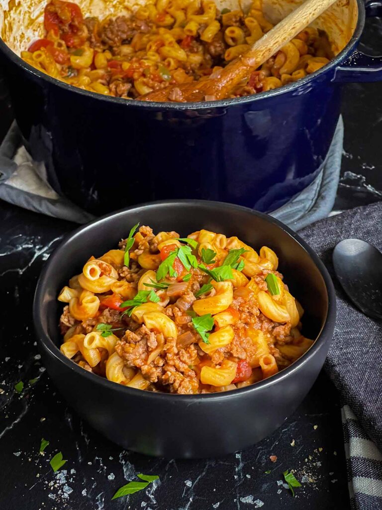 A bowl of american goulash in a dark bowl with a dutch oven full of goulash in the background.