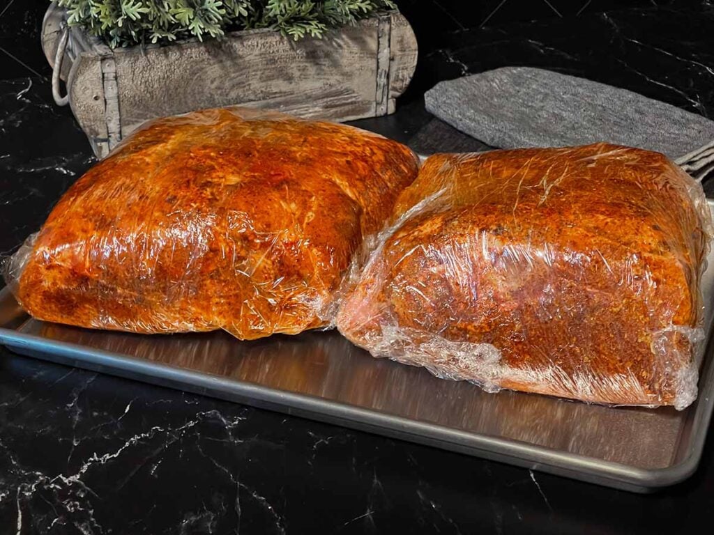 Two pork butts with rub applied, wrapped in plastic wrap.