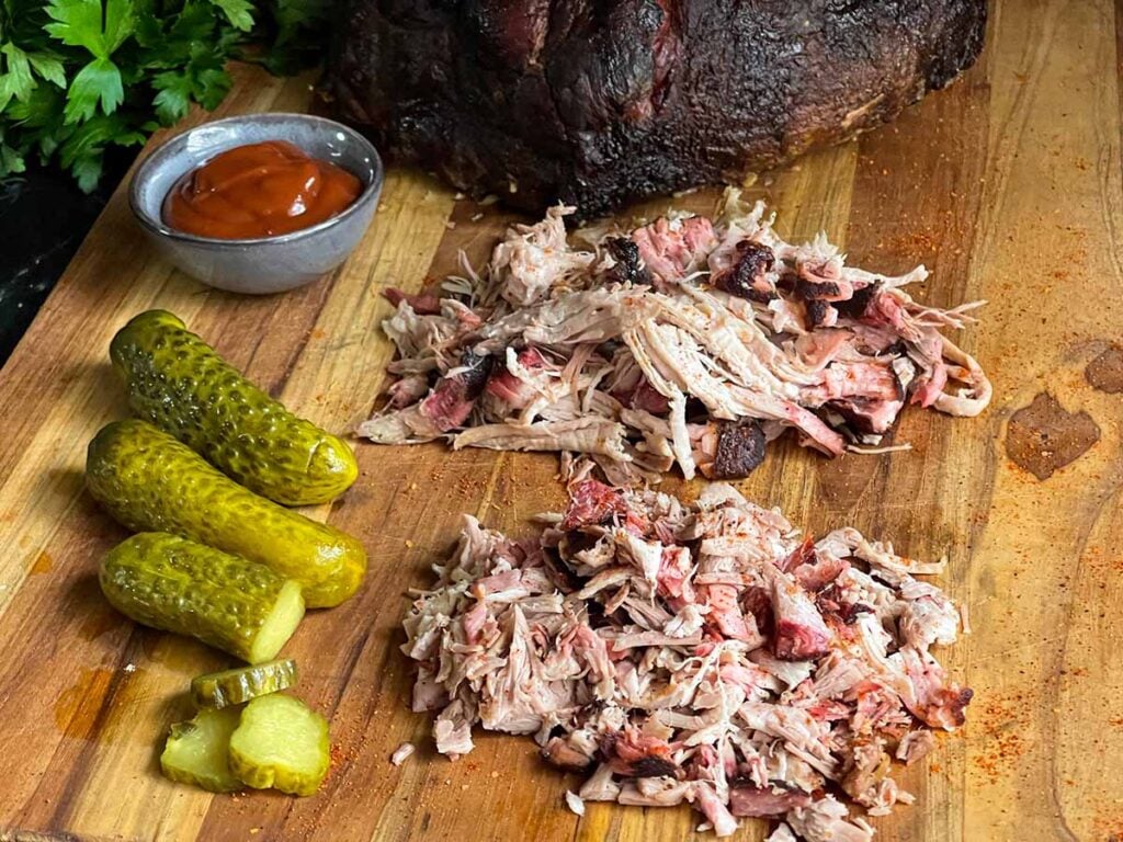 Chopped and shredded smoked pork butt on a cutting board with pickles and barbecue sauce.