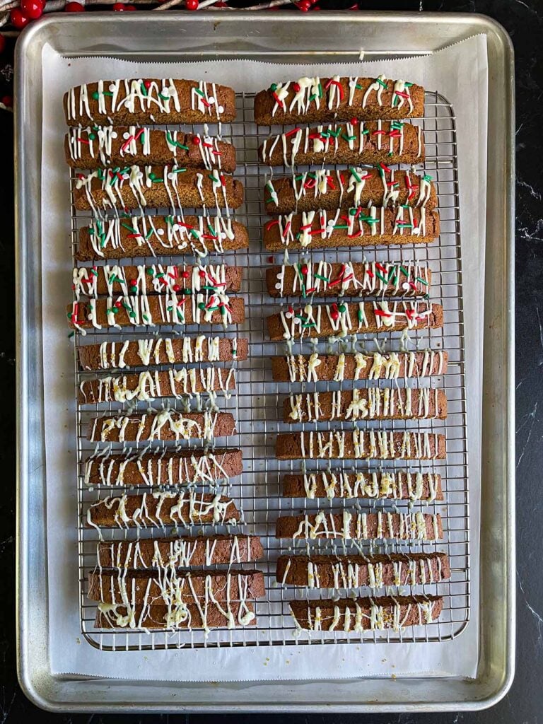 Gingerbread biscotti on a wire rack in a baking sheet drizzled with white chocolate and sprinkles.