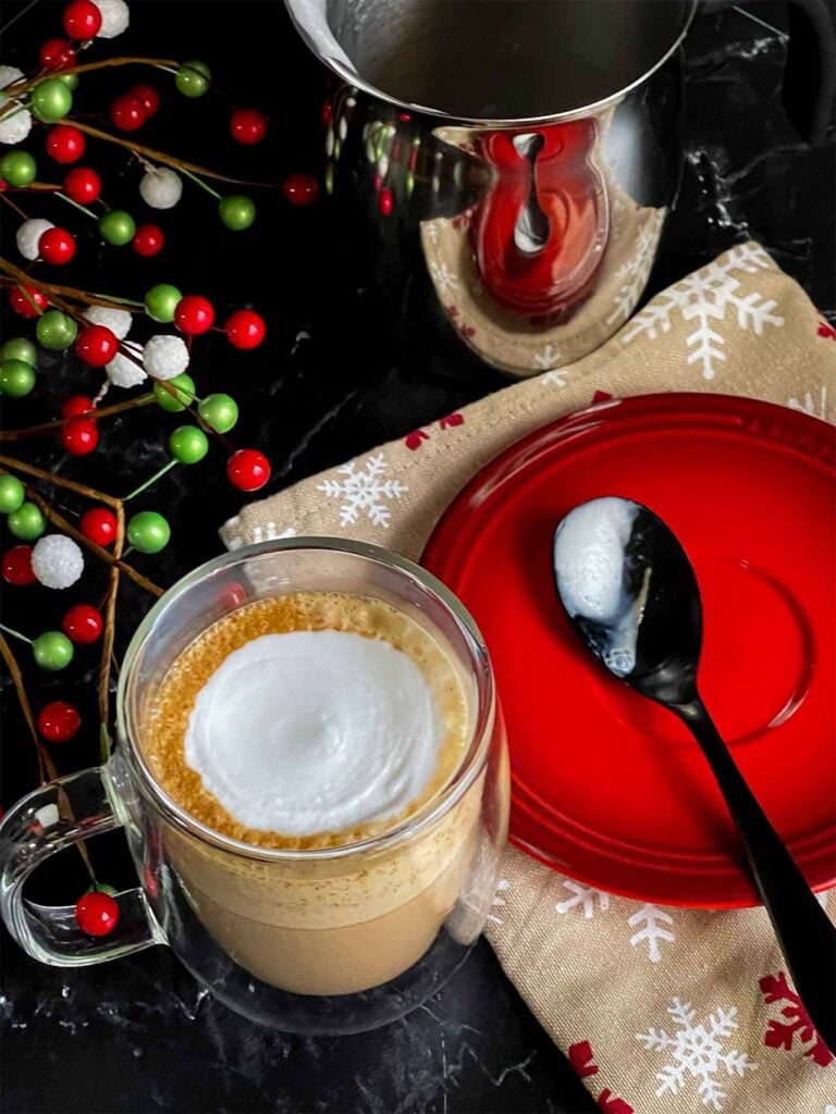 Holiday spice flat white in a glass mug. A spoon resting in a small red plate to the side.
