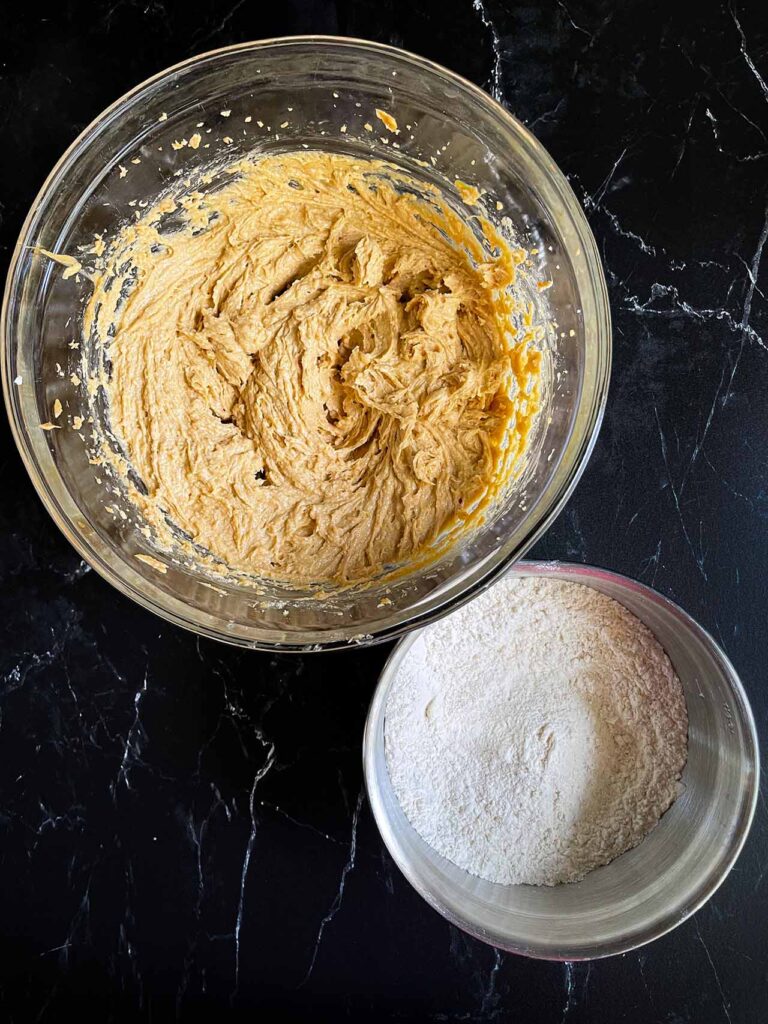 Creamed ingredients in a glass bowl and flour mixture in a metal bowl on a dark surface.