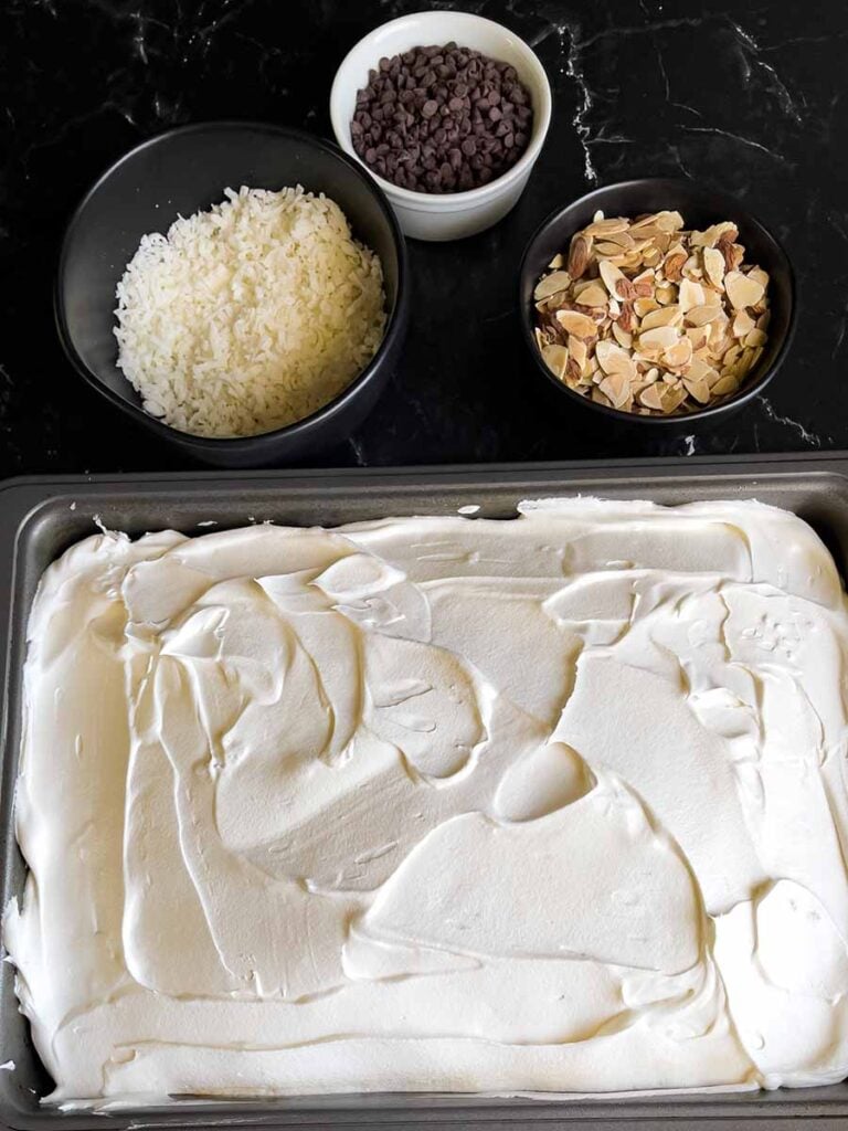 Whipped topping spread over the Almond Joy cake with coconut, almonds, and chocolate chips in the background.