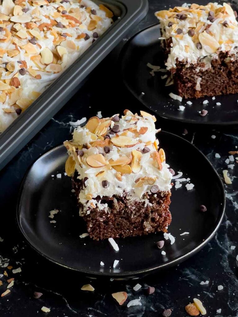 A slice of Almond Joy cake on a dark plate with the whole cake in the background.