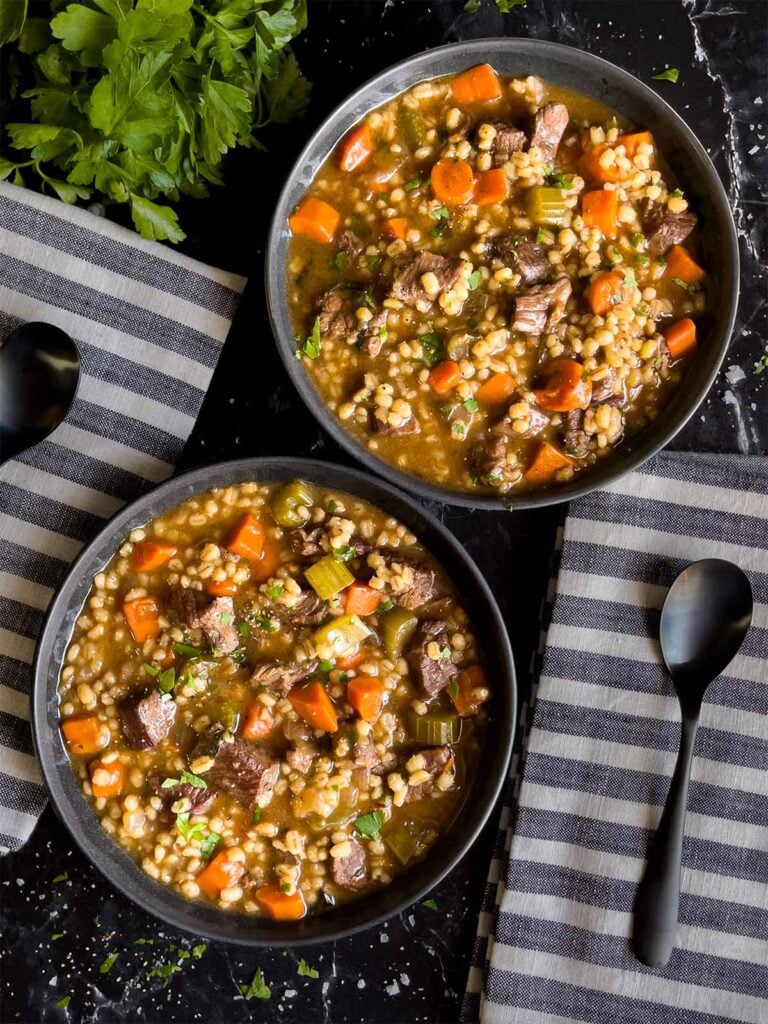 Beef barley soup in two dark bowls with striped napkins next to them.