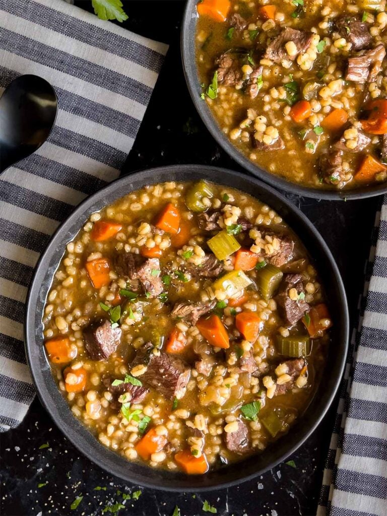 Beef barley soup in two dark bowls with striped napkins next to them.