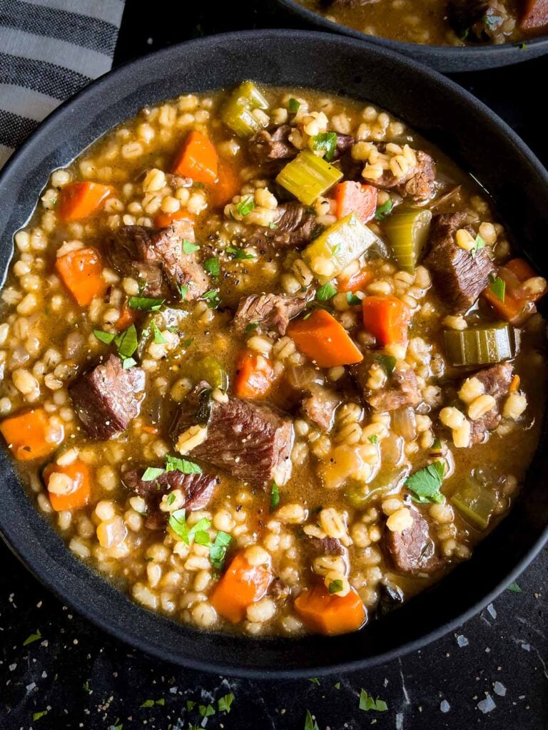 Beef barley soup in a dark bowl garnished with chopped parsley.