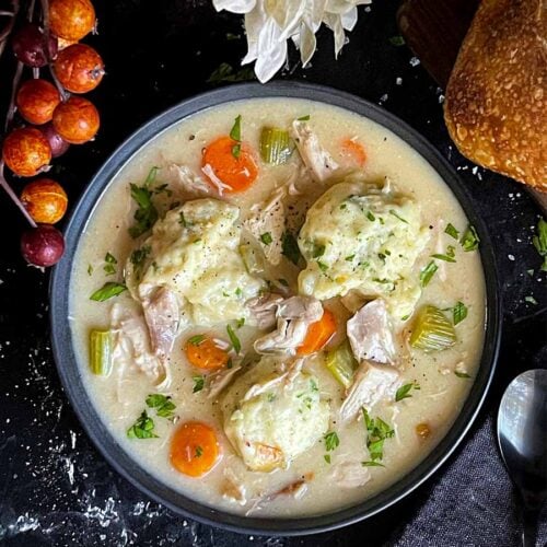 Homemade chicken and dumplings in a dark bowl with a loaf of bread in the background.